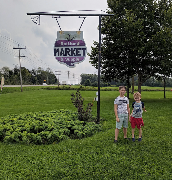 Two children in a green field with some trees in the background standing by the sign for Maitland Market and Supply in Goderich